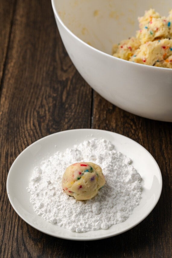 A funfetti cookie dough ball in a small bowl of powdered sugar, next to a large bowl of cookie dough.