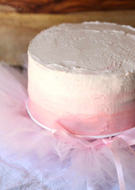 A frosted Pink Lemonade Chiffon Cake on a cake stand surrounded by tufts of pink chiffon.