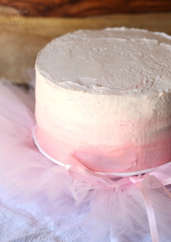 A frosted pink lemonade chiffon cake on a cake stand surrounded by clusters of pink chiffon.