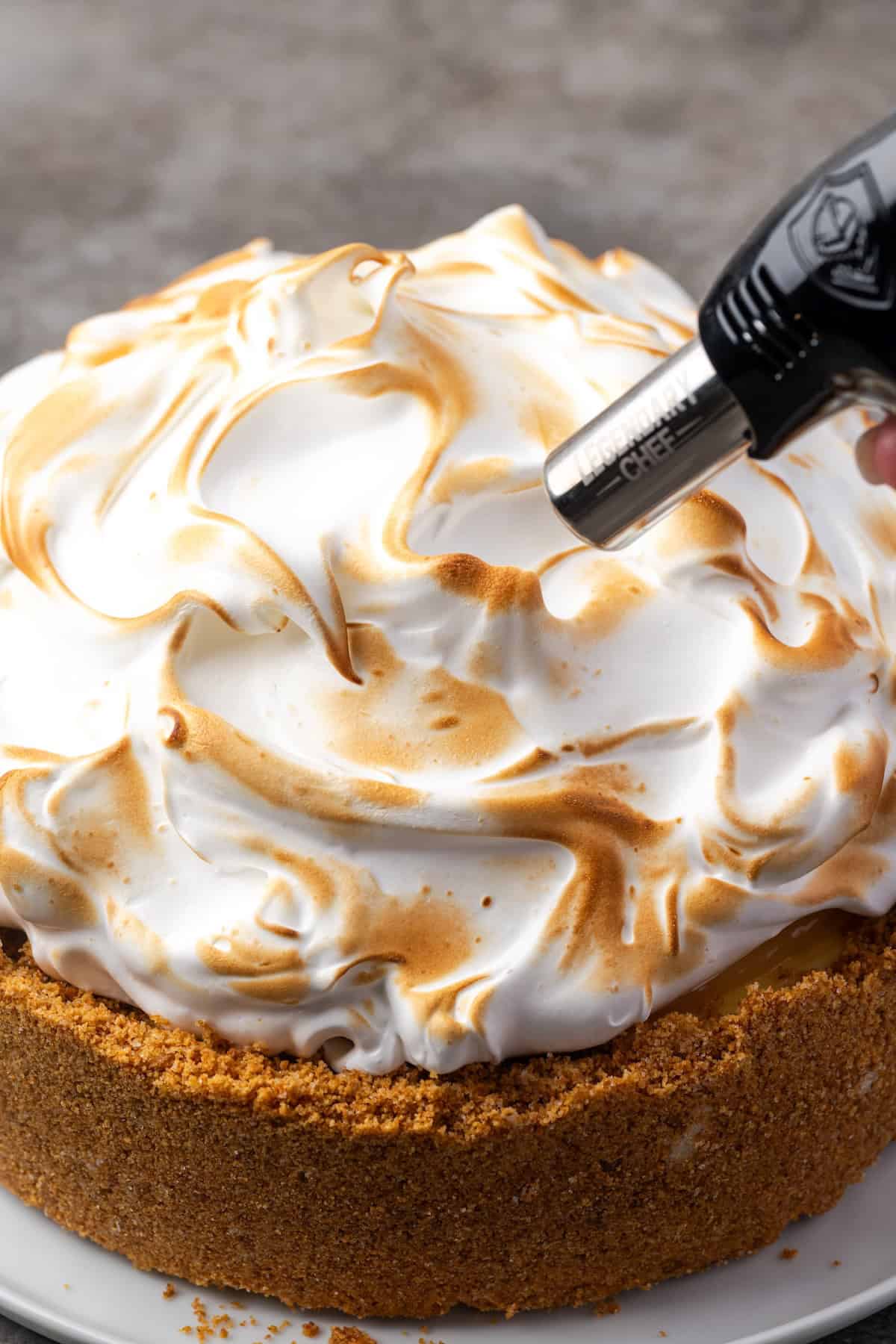 This zesty lemon meringue cheesecake has a smooth and creamy lemon filling, buttery graham cracker crust, and pillows of homemade meringue! A kitchen torch being used to toast the tips of the meringue on top of a lemon meringue cheesecake.