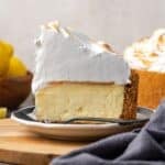 Side view of a slice of lemon meringue cheesecake on a plate with the rest of the cheesecake in the background.