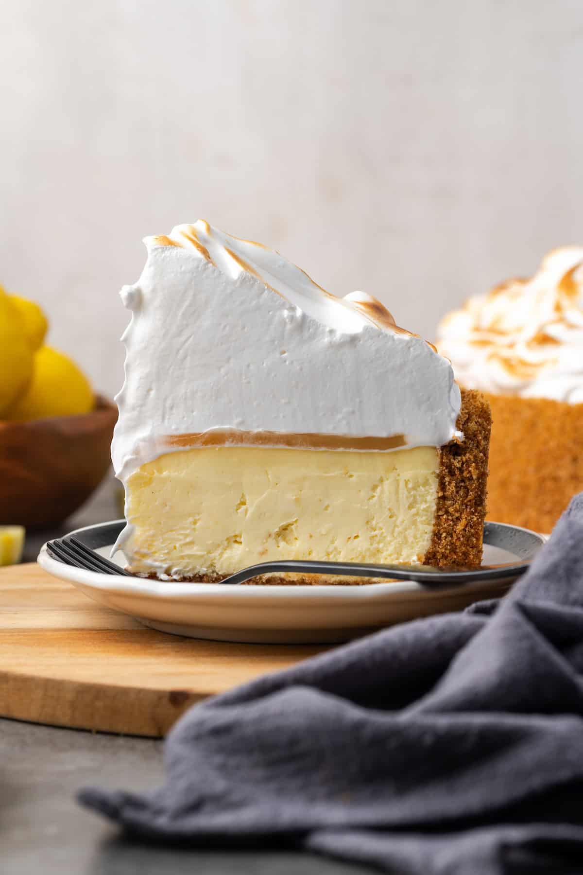 This zesty lemon meringue cheesecake has a smooth and creamy lemon filling, buttery graham cracker crust, and pillows of homemade meringue! Side view of a slice of lemon meringue cheesecake on a plate with the rest of the cheesecake in the background.