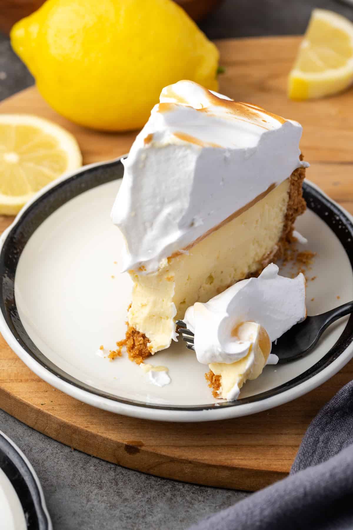 This zesty lemon meringue cheesecake has a smooth and creamy lemon filling, buttery graham cracker crust, and pillows of homemade meringue! A slice of lemon meringue cheesecake on a plate with a forkful missing from the end, and lemons sliced in the background.