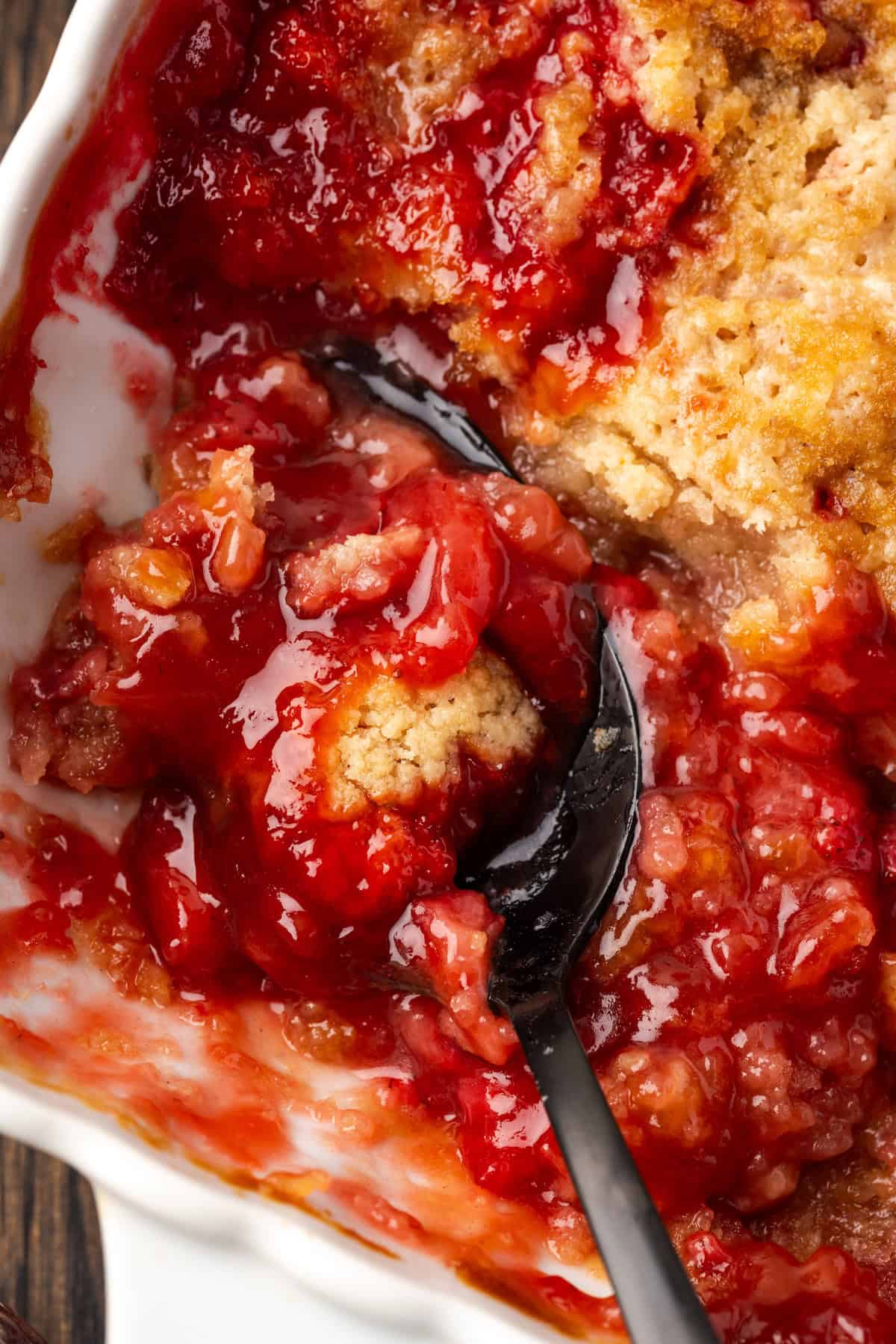 Close up of a spoon resting in an open serving of strawberry cobbler in a baking dish.