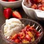 A serving of strawberry cobbler in a bowl topped with vanilla ice cream, with the rest of the cobbler in a baking dish in the background.