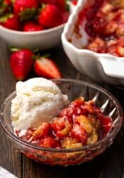 A serving of strawberry cobbler in a bowl topped with vanilla ice cream, with the rest of the cobbler in a baking dish in the background.