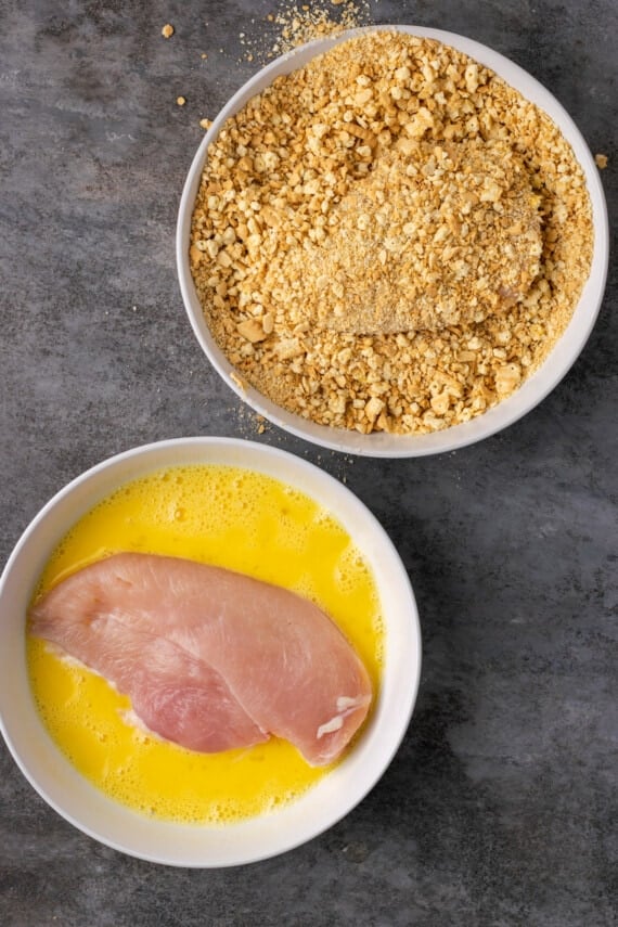 Chicken breasts in a bowl of beaten eggs, and a bowl of cracker crumbs.