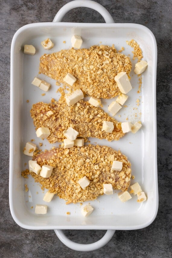 Coated chicken breasts in a baking dish with cubes of butter.