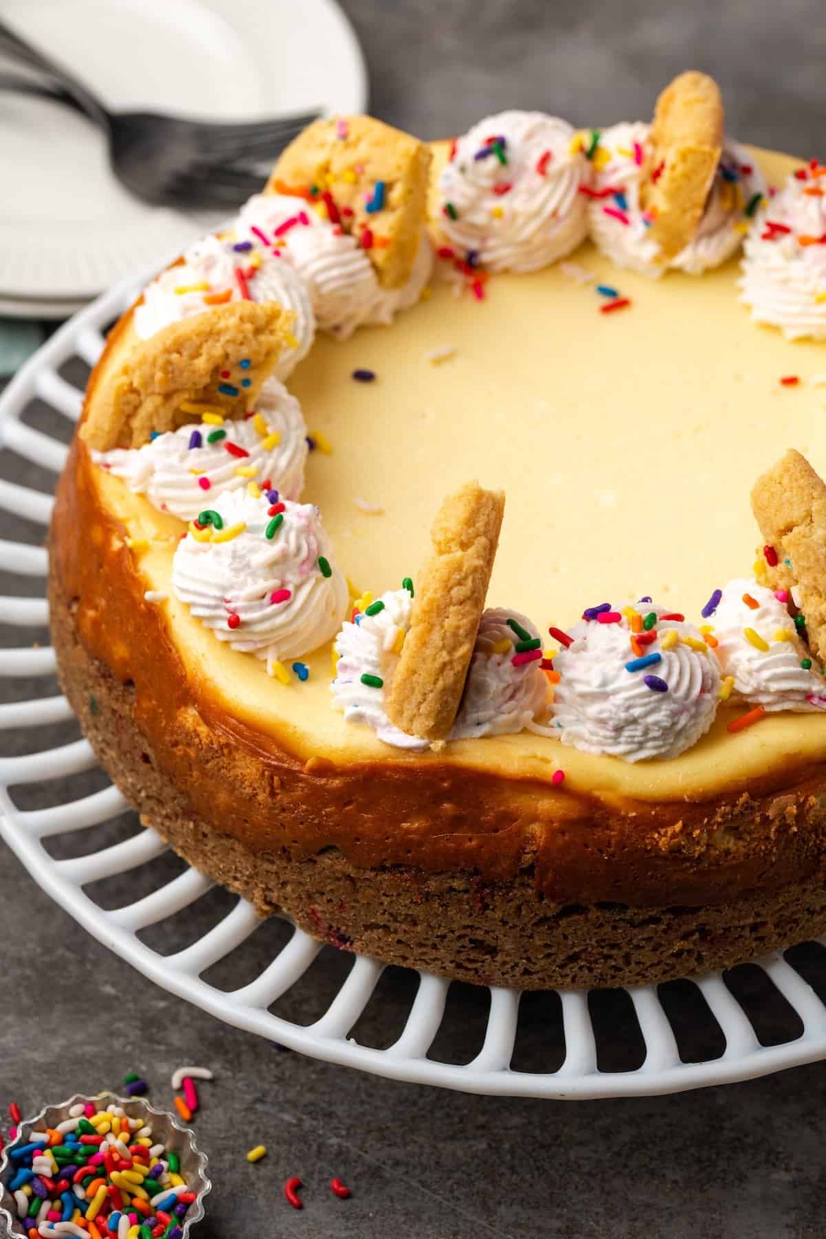 This sprinkle sugar cookie cheesecake is a creamy vanilla cheesecake baked on a buttery sugar cookie crust, topped with fluffy whipped cream! Sugar cookie cheesecake topped with swirls of sprinkle whipped cream and broken cookies.