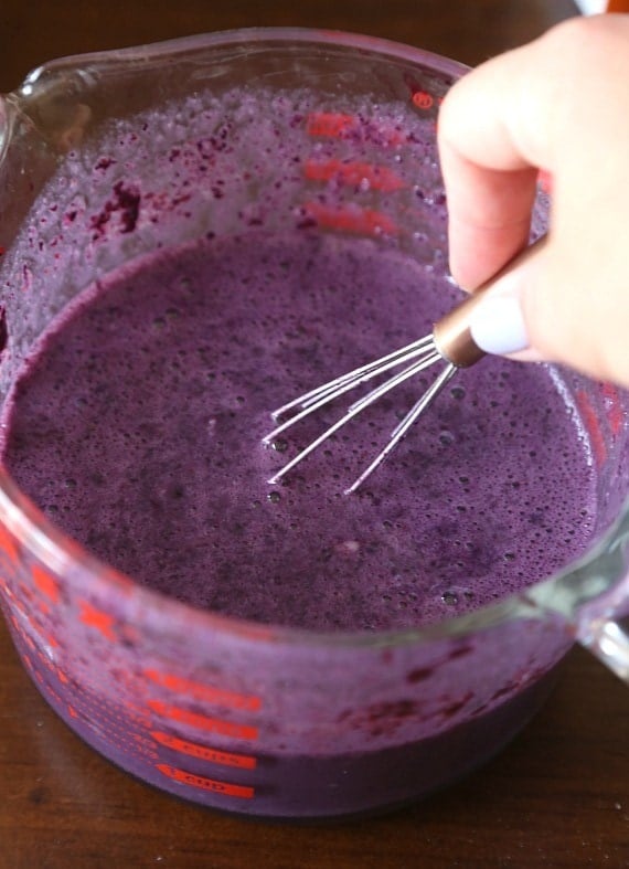 A hand using a whisk to stir blueberry puree in a measuring cup.