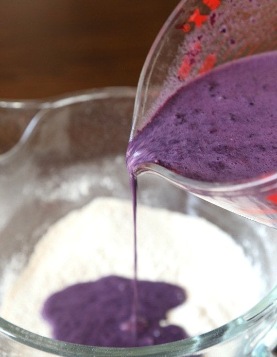 Pureed blueberries being poured from a measuring cup into a bowl of dry ingredients.