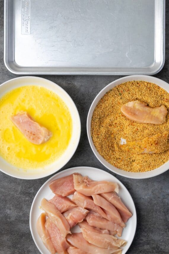 Overhead view of a chicken breading station with a bowl of chicken, stuffing crumbs, and an egg wash next to a baking pan.