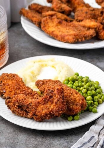 Crispy chicken tenders breaded with chicken stuffing next to mashed potatoes and peas on a plate, with a platter of chicken tenders in the background.