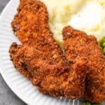 Close up of crispy chicken tenders breaded with chicken stuffing next to mashed potatoes and peas on a plate.