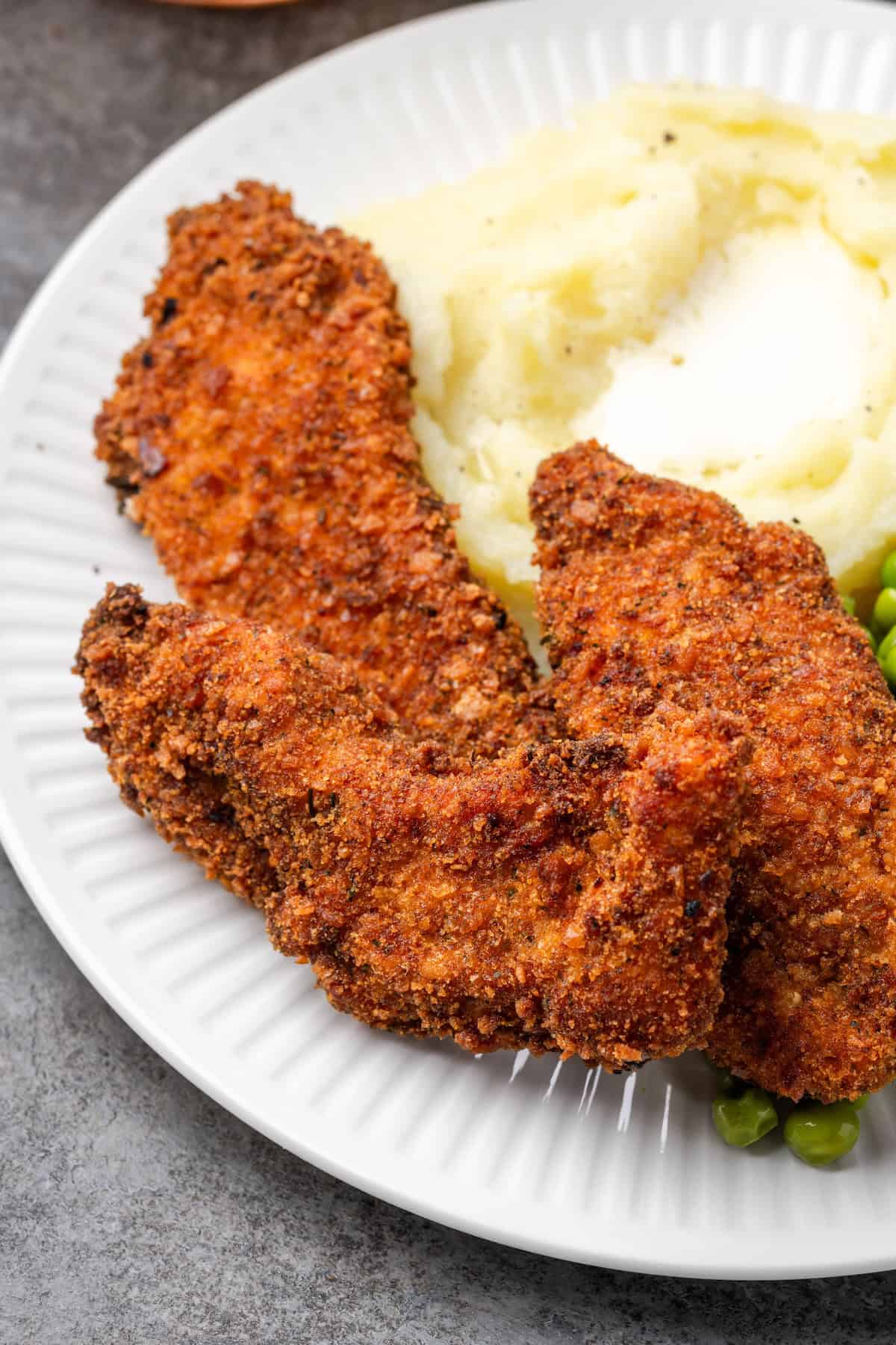 Close up of crispy chicken tenders breaded with chicken stuffing next to mashed potatoes and peas on a plate.