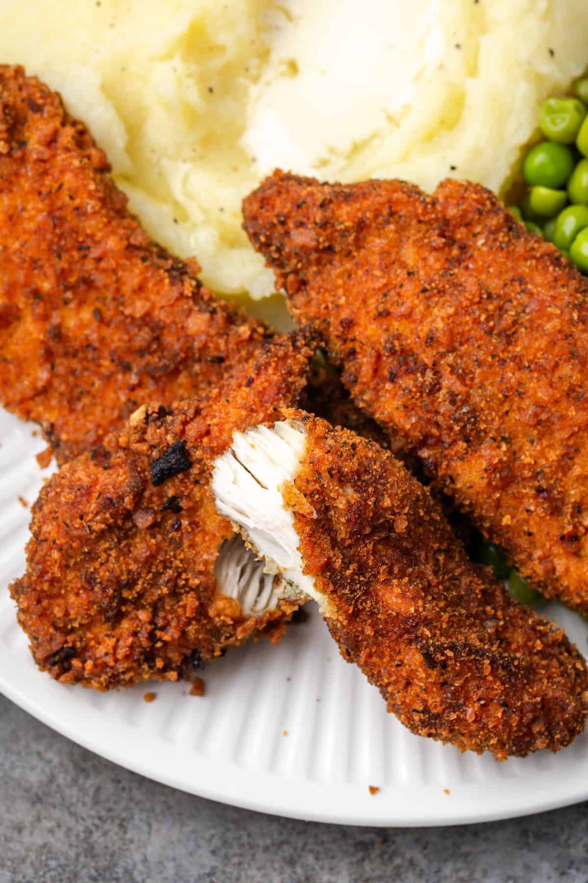 A crispy chicken tender broken in half next to mashed potatoes and peas on a plate.