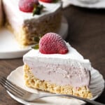 A slice of Krispie Treat ice cream pie on a white plate next to a fork, garnished with a fresh strawberry, with the rest of the pie in the background.