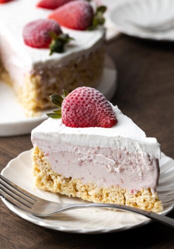 A slice of Krispie Treat ice cream pie on a white plate next to a fork, garnished with a fresh strawberry, with the rest of the pie in the background.