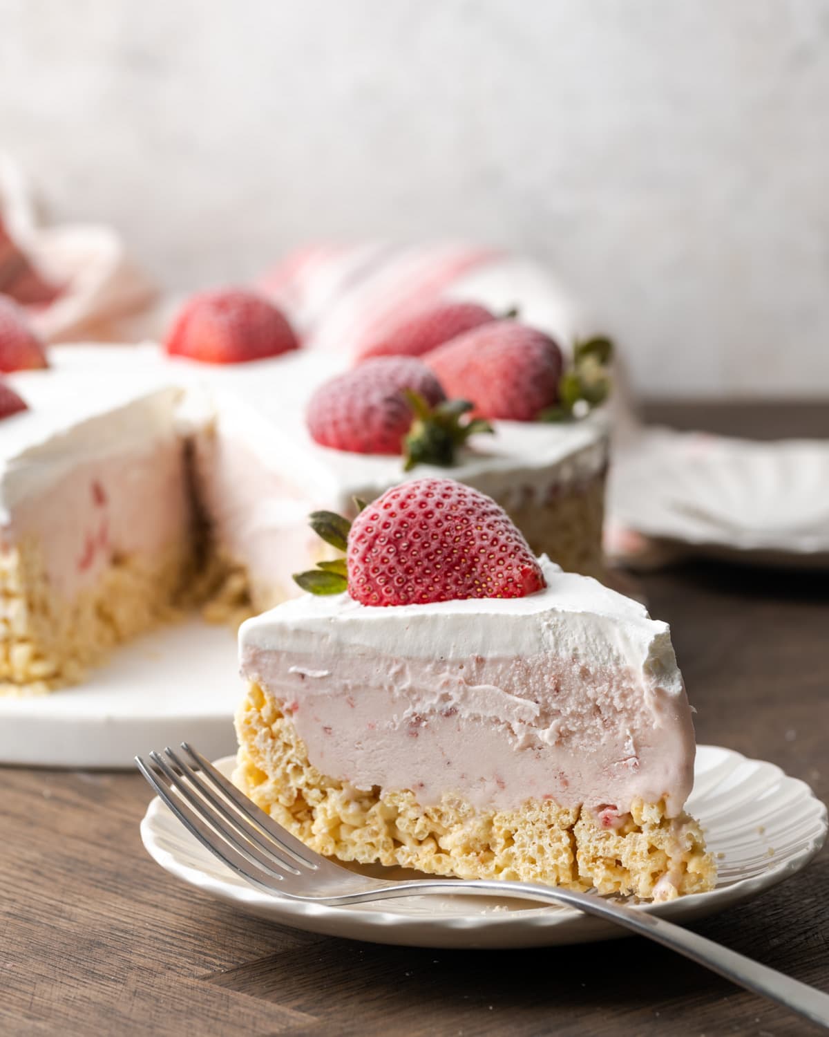 A slice of Krispie Treat ice cream pie on a white plate next to a fork, garnished with a fresh strawberry, with the rest of the pie in the background.