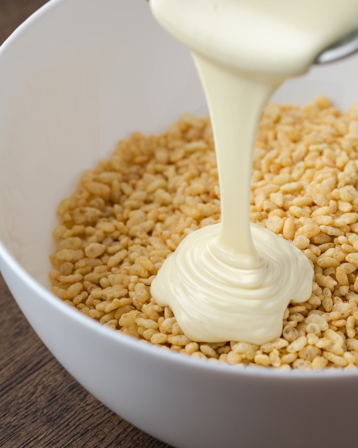 Melted white chocolate is poured into a white mixing bowl with Rice Krispie cereal.
