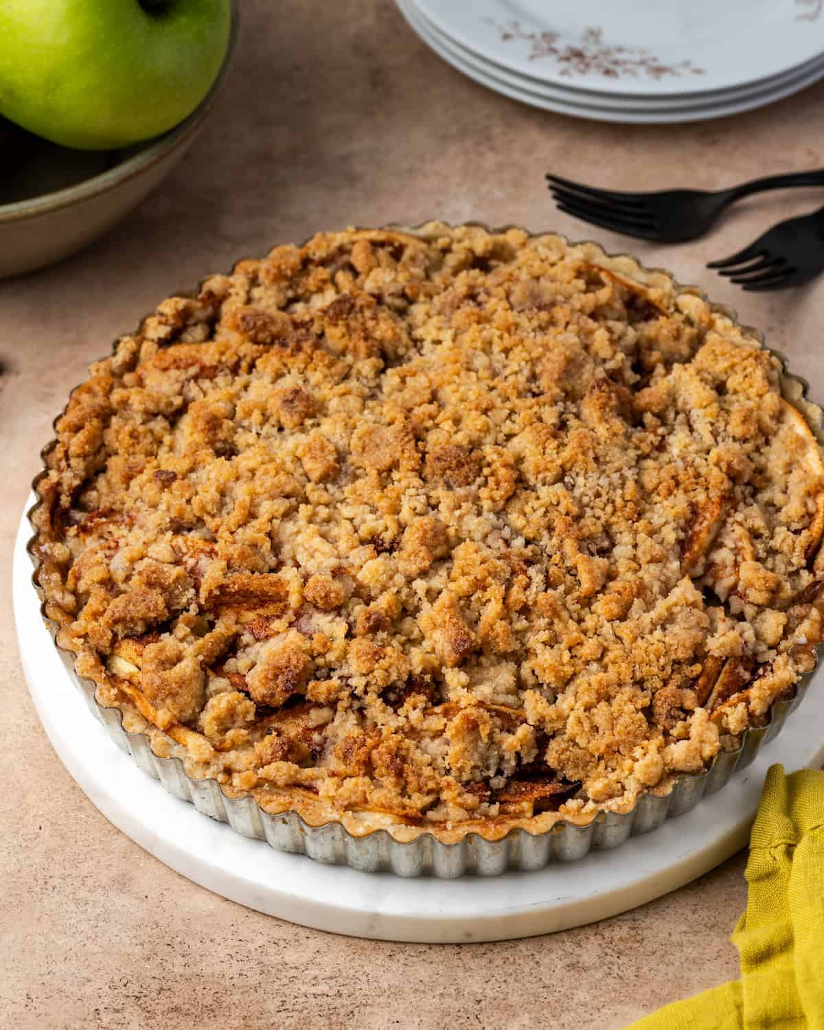 Baked apple crumble tart in a tart pan with a stack of plates and a bowl of apples in the background.