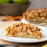 A slice of apple crumble tart on a plate with a bowl of apples and the rest of the tart in the background.