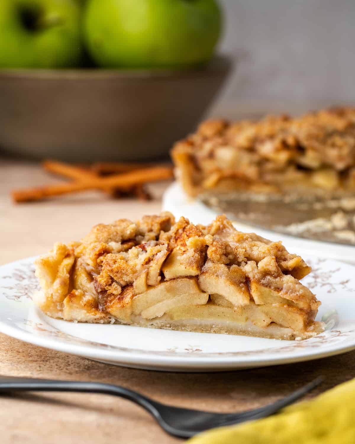 A slice of apple crumble tart on a plate with a bowl of apples and the rest of the tart in the background.