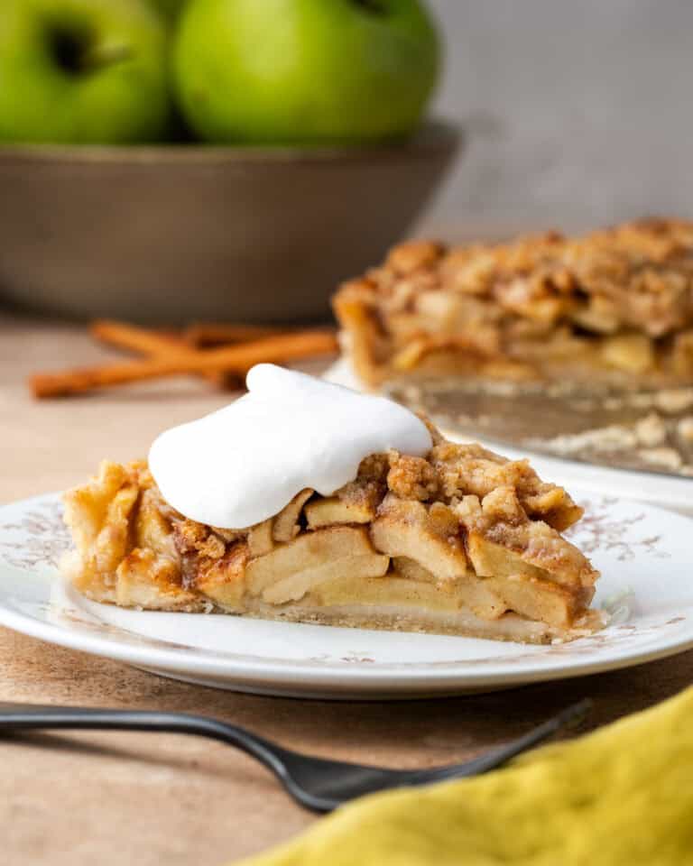 A slice of apple crumble tart topped with whipped cream on a plate with a bowl of apples and the rest of the tart in the background.