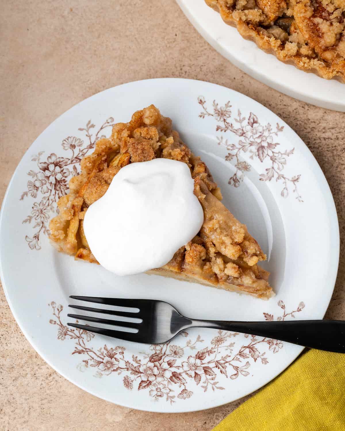 Overhead view of a slice of apple crumble tart topped with whipped cream on a plate next to a fork.