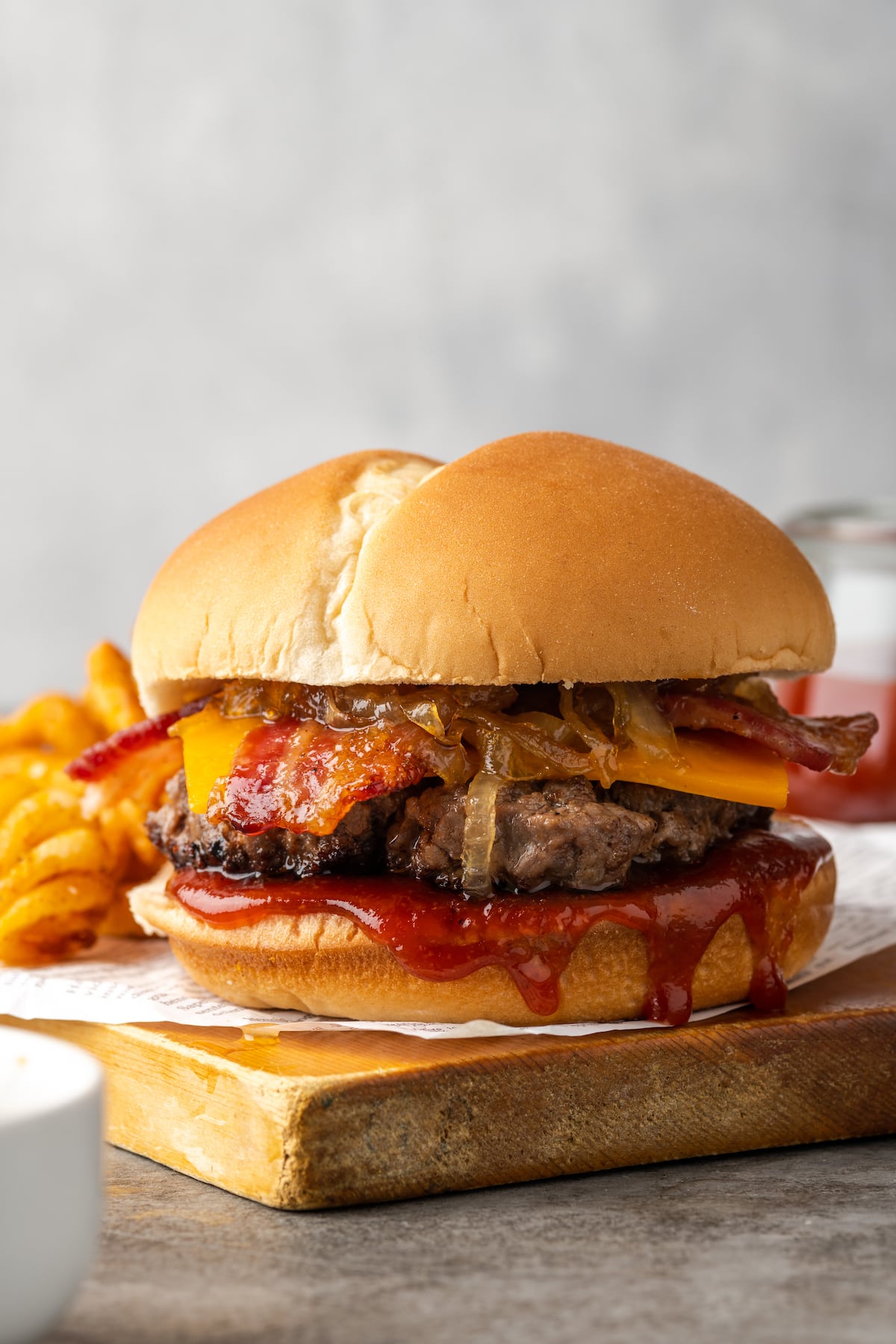 Candied bacon maple cheddar burger on a wooden board next to curly fries.