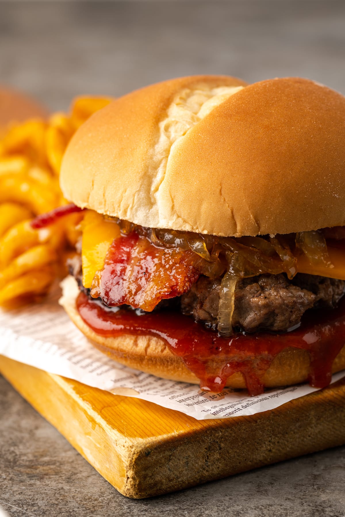 Candied bacon maple cheddar burger on a wooden board next to curly fries.