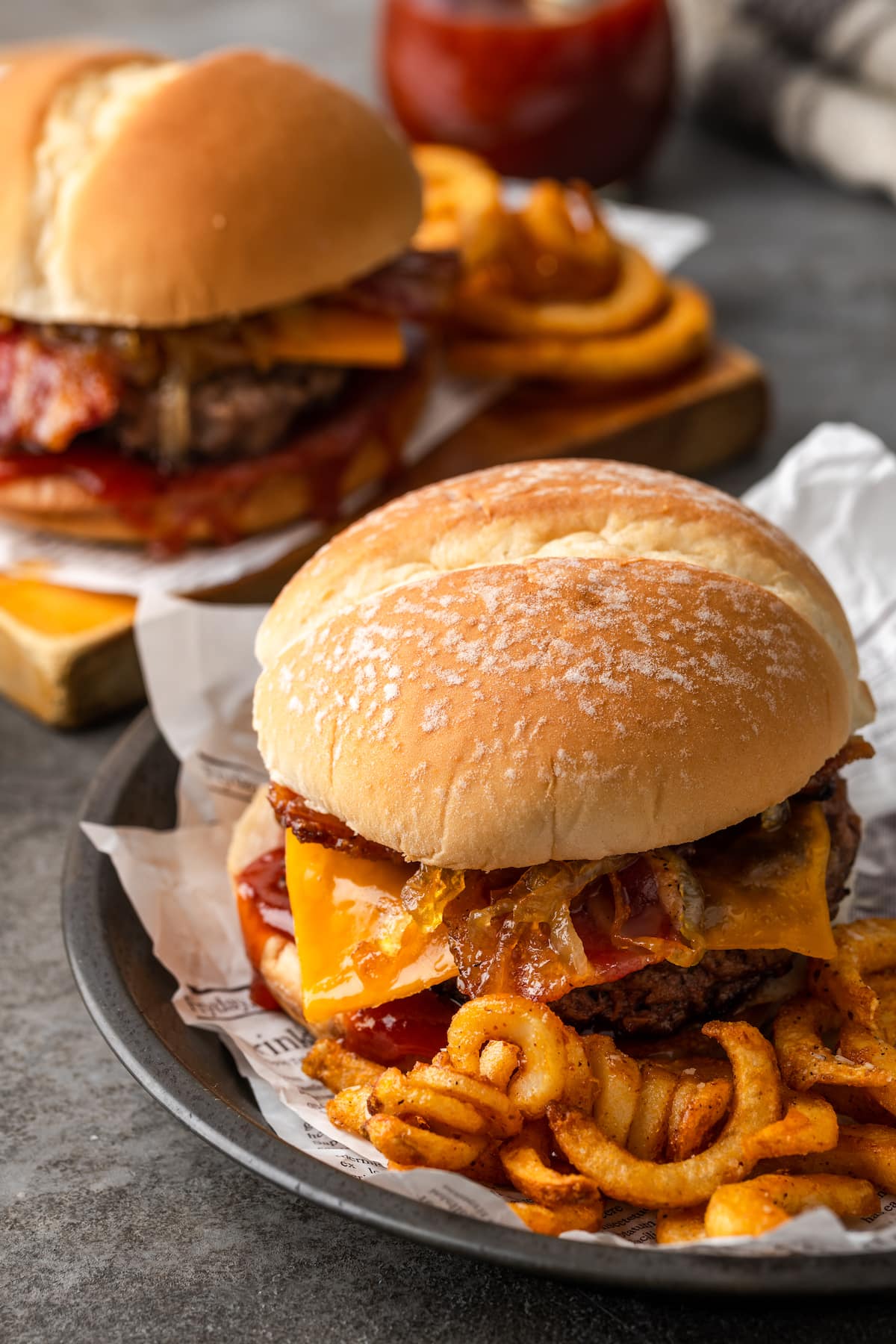 Two candied bacon maple cheddar burgers served on plates with curly fries.