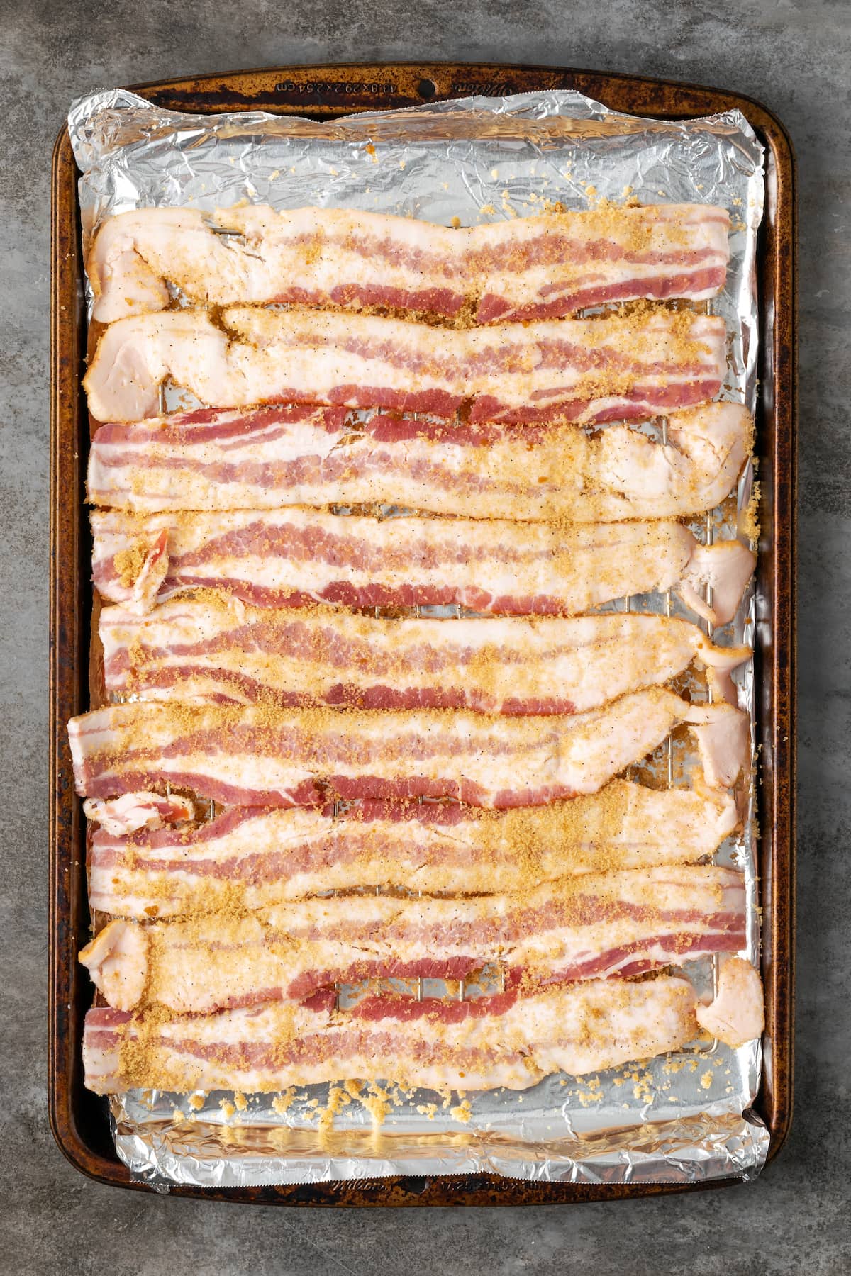 Overhead view of strips of raw bacon lined up on a baking sheet sprinkled with brown sugar.