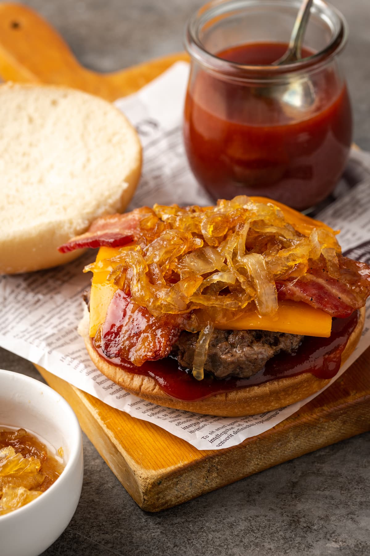 A partially assembled candied bacon maple cheddar burger topped with caramelized onions on a wooden board next to a jar of BBQ sauce.