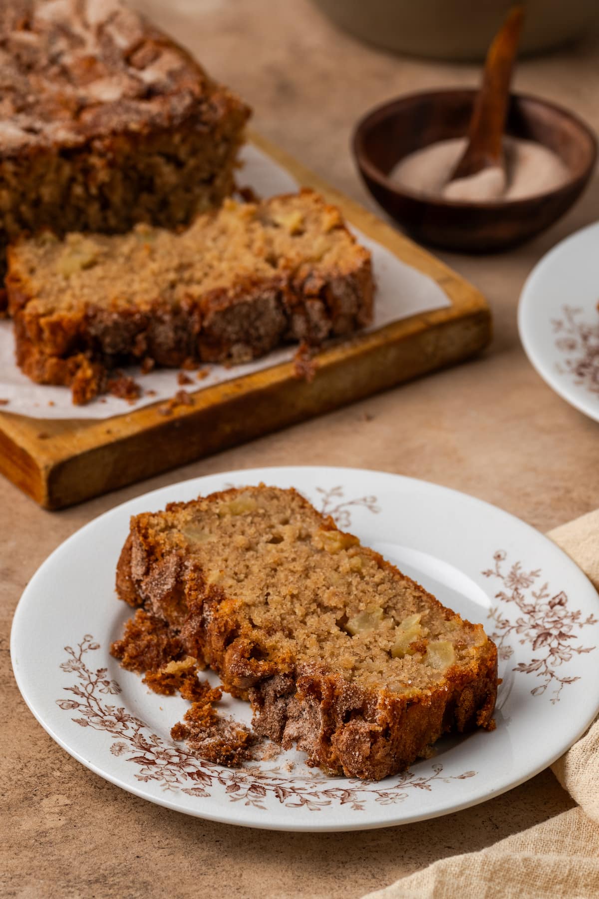 A slice of apple cinnamon bread on a white plate with more bread slices on a wooden cutting board in the background.