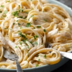 A bowl of fettuccine Alfredo garnished with fresh chopped parsley, with silver serving utensils.