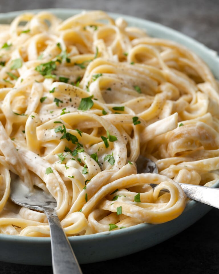 A bowl of fettuccine Alfredo garnished with fresh chopped parsley, with silver serving utensils.