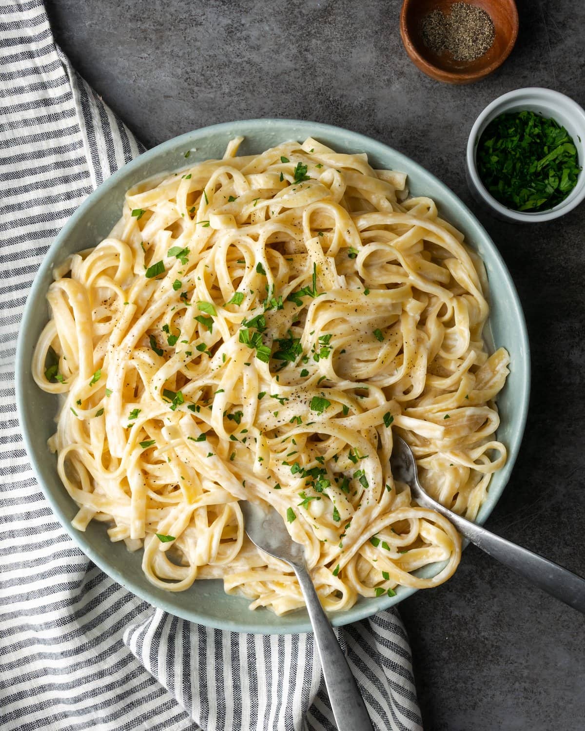 Top view of a bowl of fettuccine Alfredo garnished with fresh chopped parsley, with silver serving utensils.