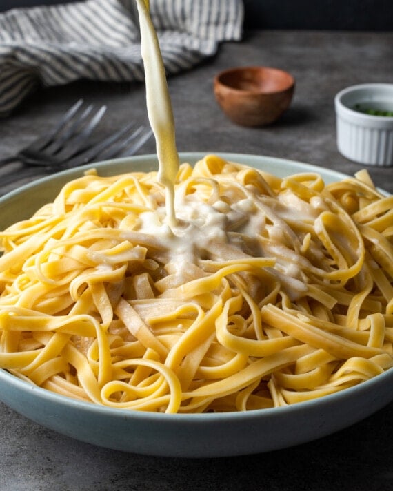 Alfredo sauce is added to a large serving bowl of cooked fettuccine pasta.
