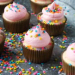 Confetti Cupcakes with pink frosting and sprinkles on top in a tray covered in sprinkles