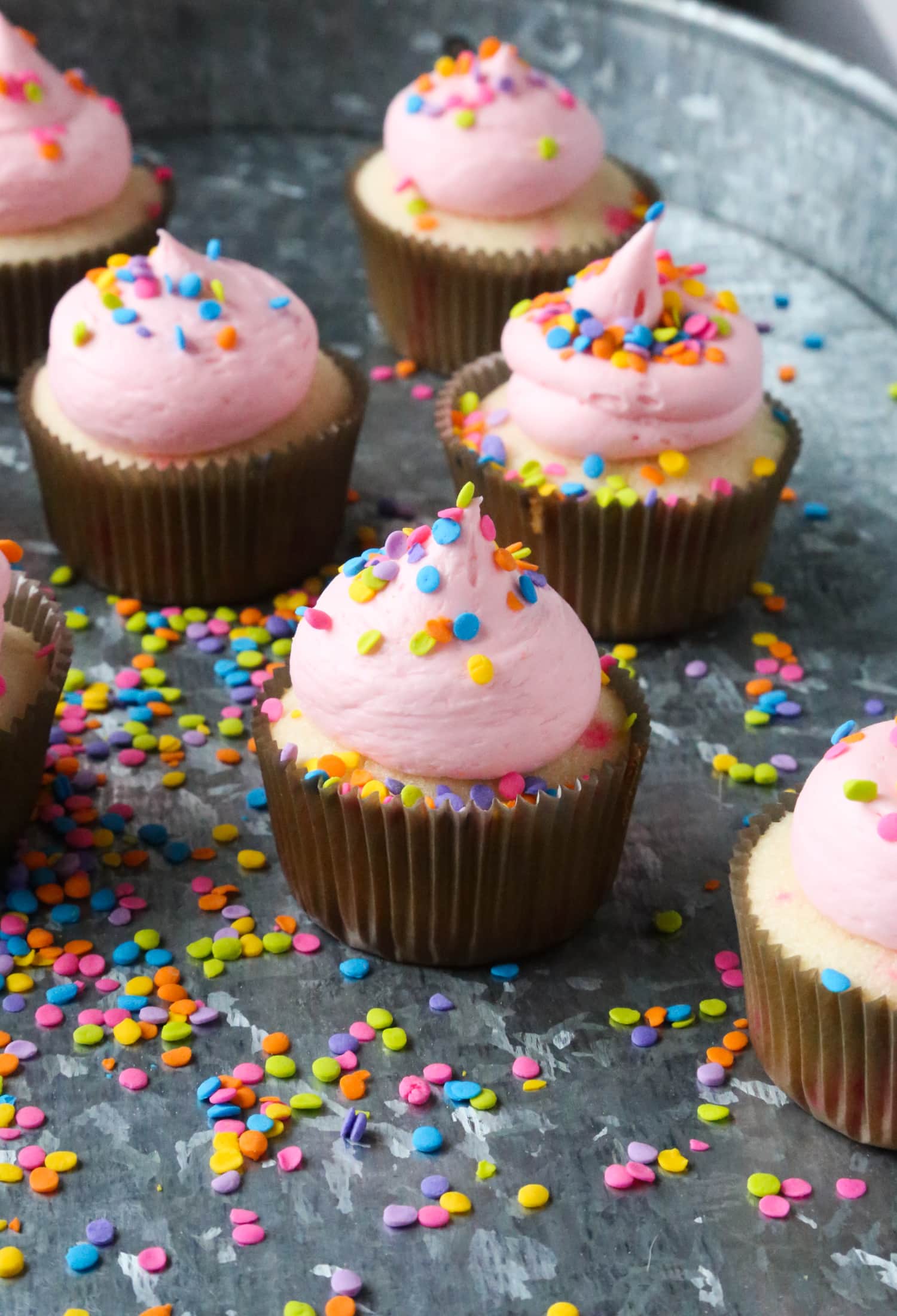 Confetti cupcakes with pink frosting and sprinkles on a sprinkle-covered tray