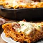 Close up of a slice of skillet lasagna on a white plate with the rest of the lasagna in a skillet in the background.