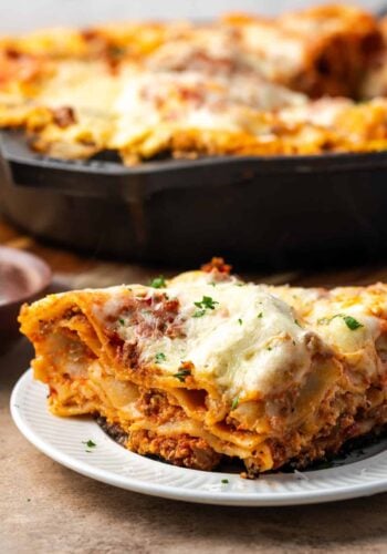 Close up of a slice of skillet lasagna on a white plate with the rest of the lasagna in a skillet in the background.