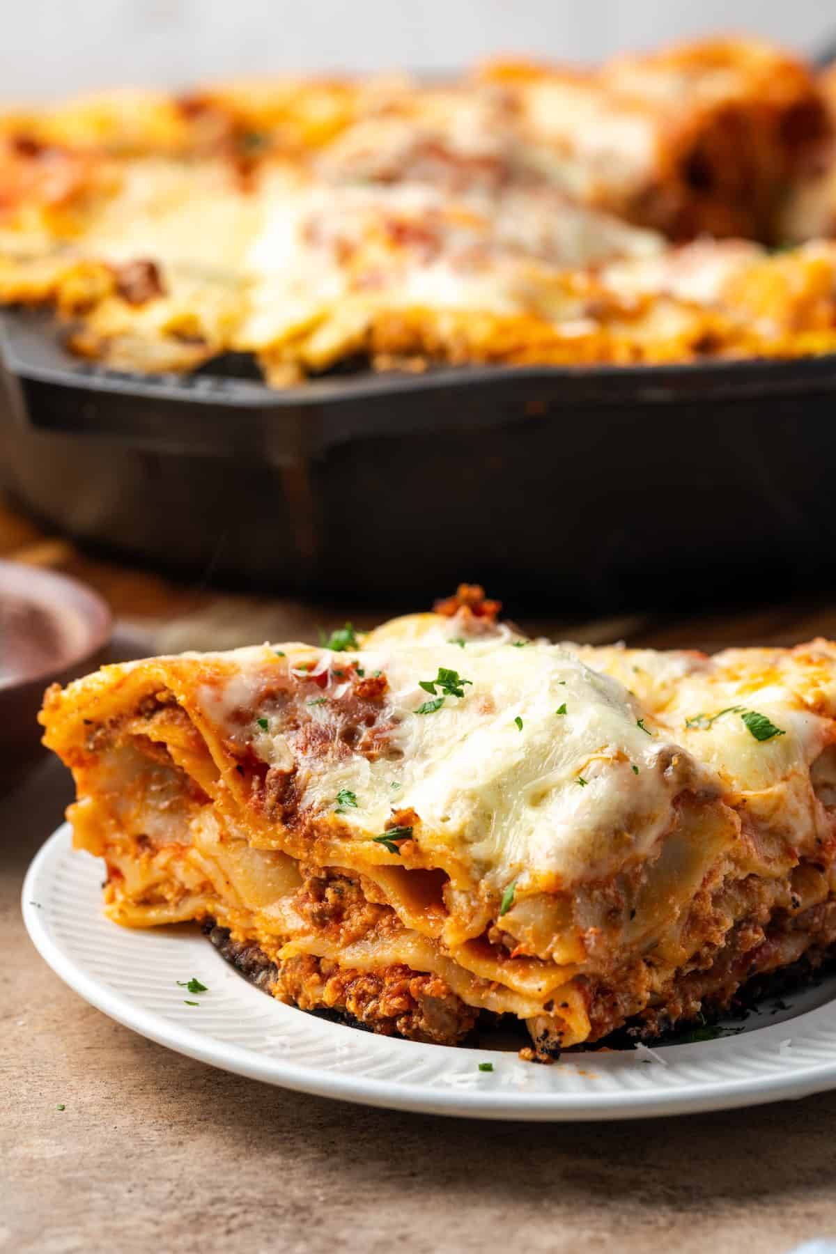 Close up of a slice of skillet lasagna on a white plate with the rest of the lasagna in a skillet in the background.
