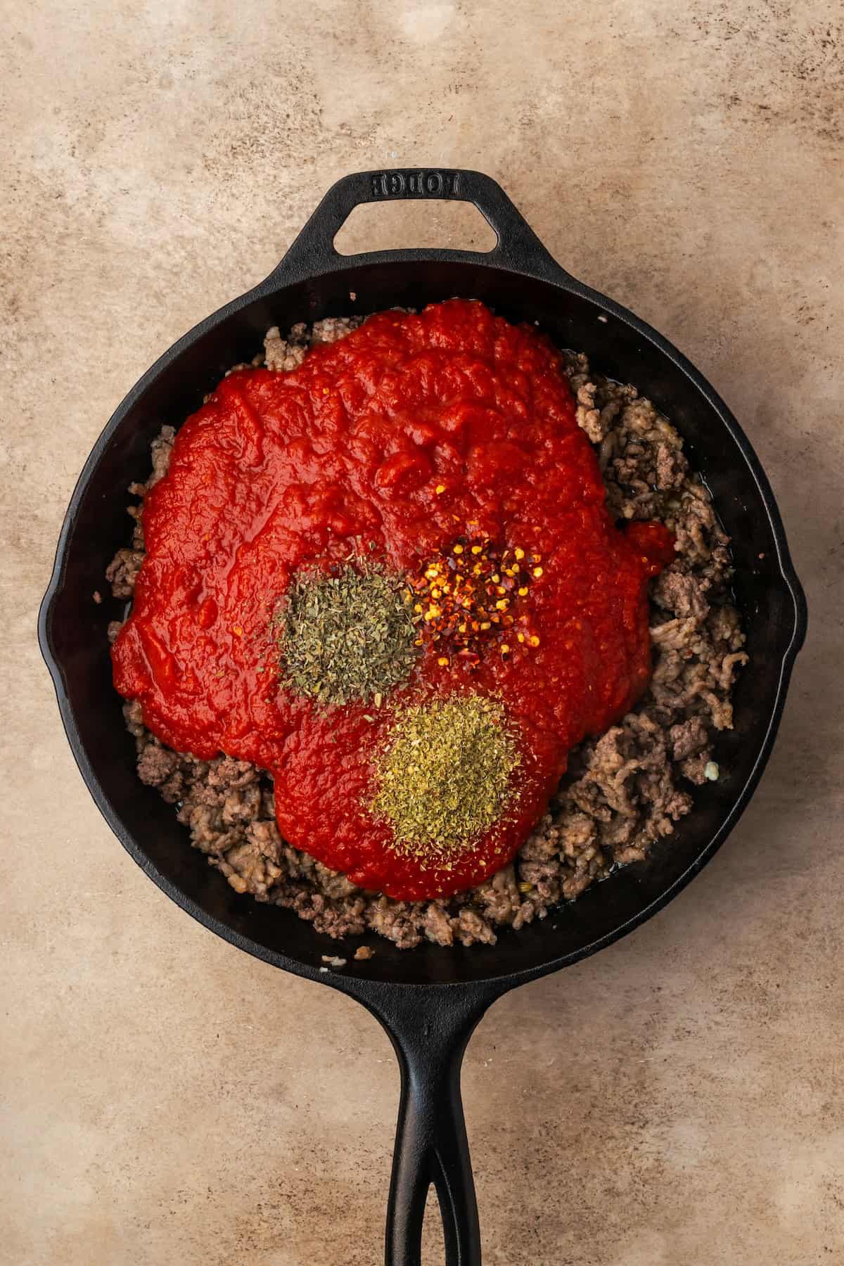 Crushed tomatoes and seasonings added to browned ground meat in a skillet.
