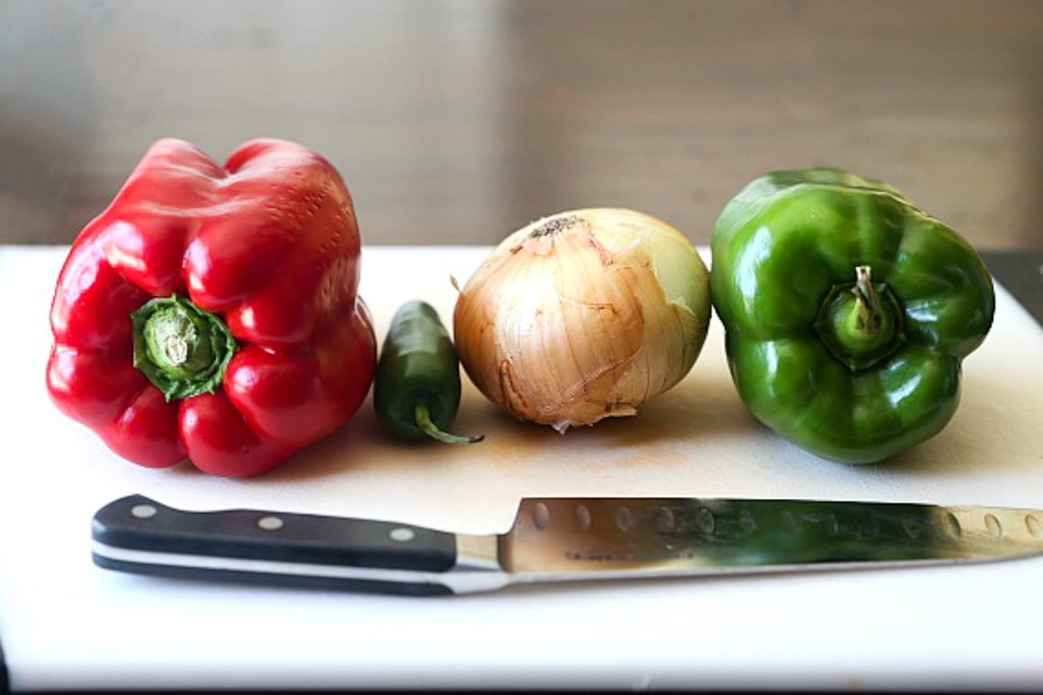 a red bell pepper, a jalapeno, a spanish onion, and a green bell pepper on a white cutting board with a chef's knife