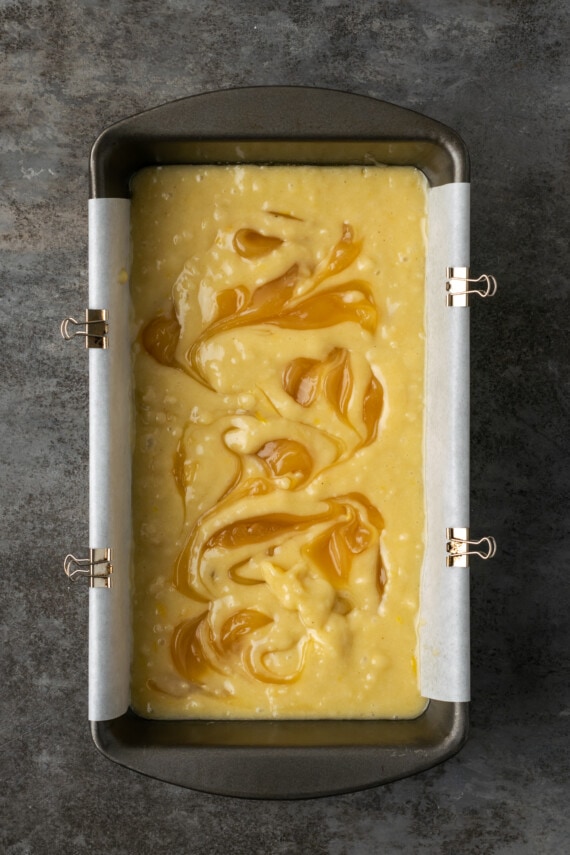 Overhead view of lemon curd swirled into lemon pound cake batter in a lined loaf pan.