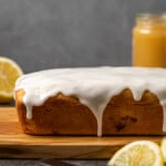 Lemon pound cake topped with lemon glaze on a cutting board, next to a jar of lemon curd and lemon wedges.