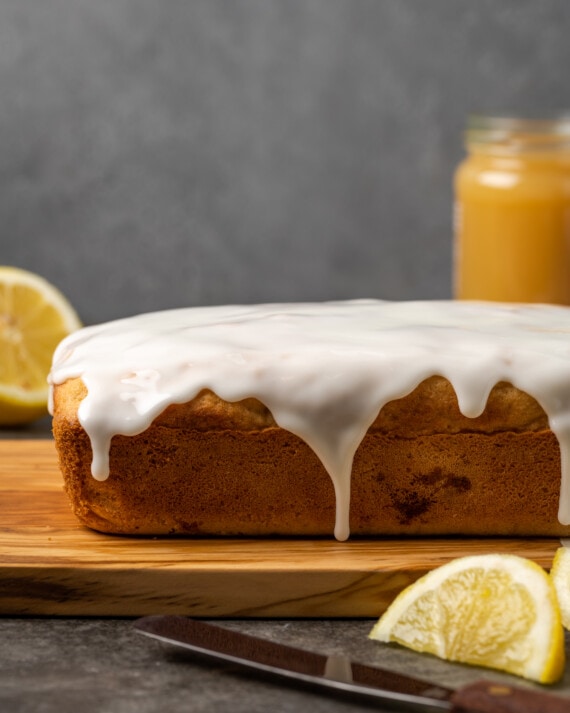 Lemon pound cake topped with lemon glaze on a cutting board, next to a jar of lemon curd and lemon wedges.