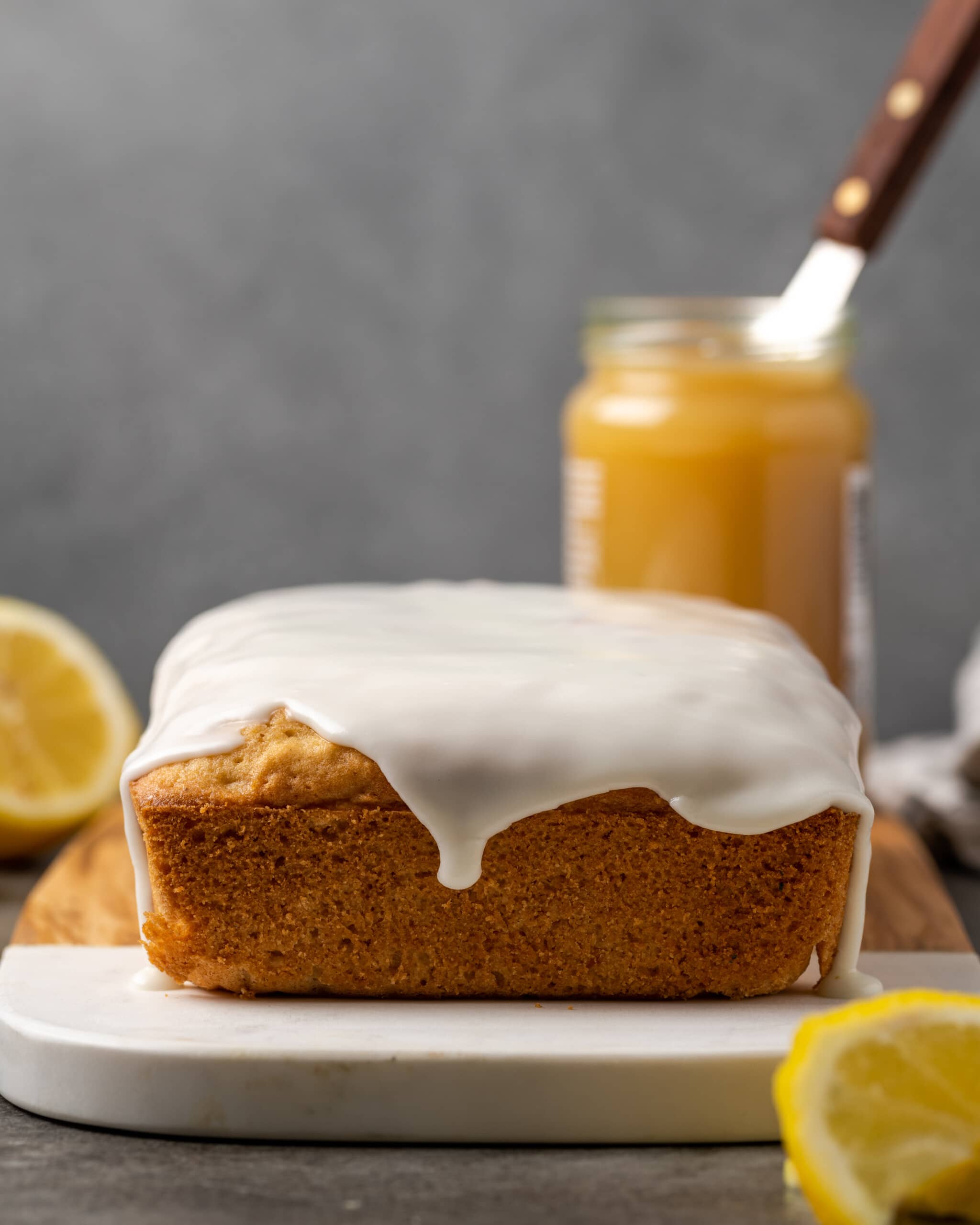 Lemon pound cake topped with lemon glaze on a cutting board, next to a jar of lemon curd and lemon wedges.